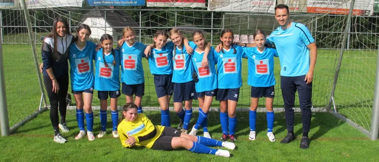 Eine Fußballmannschaft junger Mädchen in blauen Trikots steht vor einem Tor. Ein Mann steht neben ihnen und ein anderes Mädchen liegt auf dem Rasen.