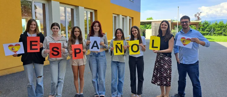 Sieben Schüler und zwei Lehrer halten Buchstaben hoch, die 'Espanol' buchstabieren, vor einem bunten Gebäude. Sie lächeln und posieren für ein Foto.