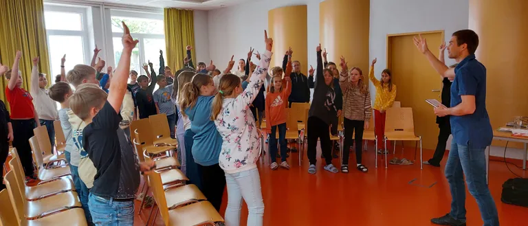 Eine Gruppe von Kindern und einigen Erwachsenen steht in einem Klassenzimmer mit orangem Boden und hebt die Hände. Ein Ausbilder steht davor.