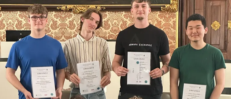 Sieben junge Männer posieren mit Urkunden in der Hand für ein Foto. Sie alle lächeln und stehen vor einer dekorativen Wand. Ein Mann trägt ein schwarzes Armani Exchange-Shirt.