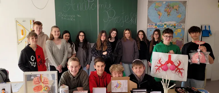 Eine Gruppe von Schülern posiert für ein Foto im Klassenzimmer. Sie stehen vor einem Tisch mit verschiedenen Gegenständen, darunter ein Gehirnmodell, ein Plakat einer Lunge und eine Weltkarte.