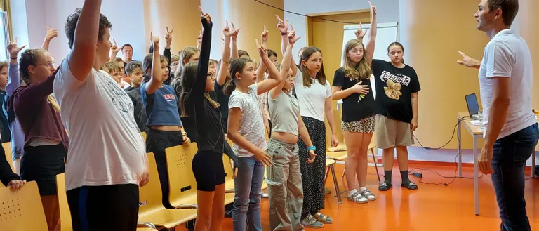 Eine Gruppe von Schülern steht und hebt die Hände in einem Klassenzimmer. Eine Projektor ist an der Decke montiert.