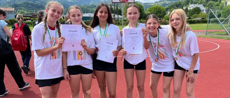 Fünf junge Mädchen mit Medaillen und Urkunden posieren für ein Foto auf einer roten Laufbahn, mit einem Basketballkorb und Bergen im Hintergrund.
