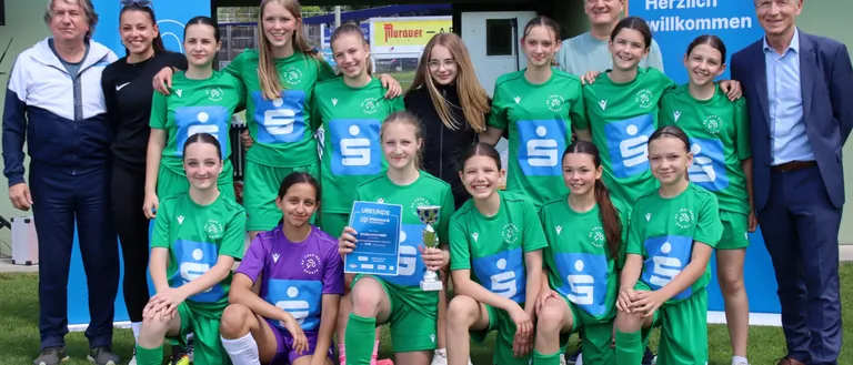 Eine Fußballmannschaft posiert mit einer Trophäe und einem Zertifikat auf einem Feld, wobei einige Personen hinter ihnen stehen und zwei Banner im Hintergrund zu sehen sind.