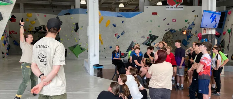Eine Gruppe von Menschen ist um eine Rednerin in einem Fitnessstudio mit einer Kletterwand versammelt. Einige Leute sitzen, während andere stehen.