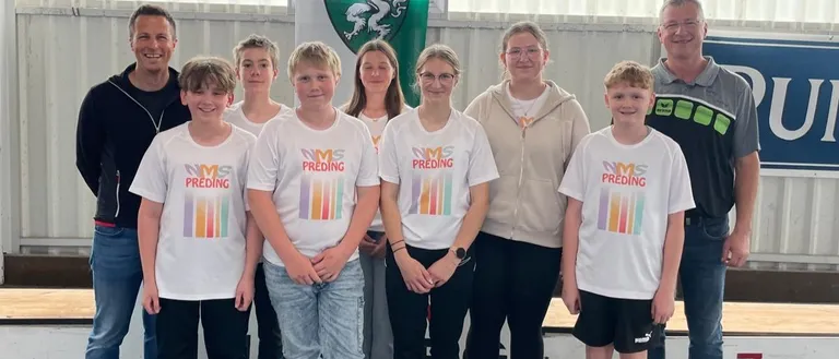 Eine Gruppe von Kindern und Erwachsenen, die weiße T-Shirts mit dem Aufdruck 'NMS Preding' tragen, steht zusammen mit Besen vor einer weißen Wand mit einem grünen Banner.