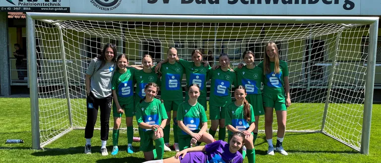 Eine Mädchenfußballmannschaft in grünen Trikots posiert vor einem Tor mit einem Gebäude namens SV Bad Schwanbe im Hintergrund.