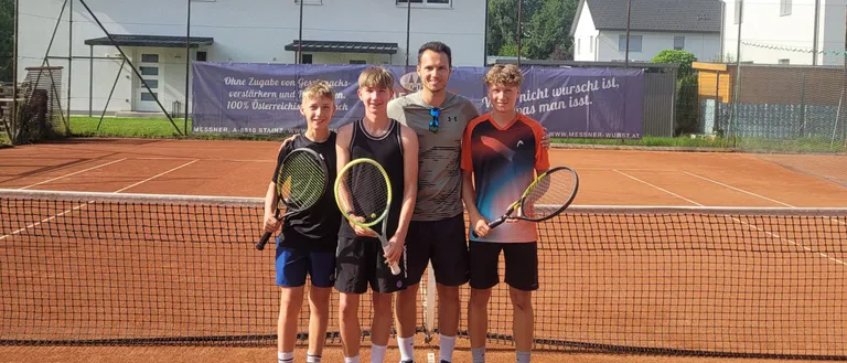 Vier Personen stehen auf einem Tennisplatz. Drei halten Tennisschläger, einer hat eine Sonnenbrille. Sie stehen vor einem Netz und einem Banner. Dahinter steht ein Haus mit einem roten Dach.
