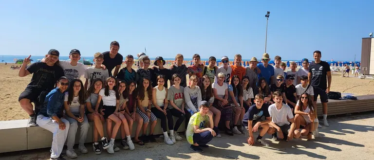Eine Gruppe von Menschen, wahrscheinlich eine Schulklasse, posiert für ein Foto an einem sonnigen Tag am Strand. Einige tragen Hüte und Sonnenbrillen. Im Hintergrund befinden sich ein Grasbereich und der Ozean.