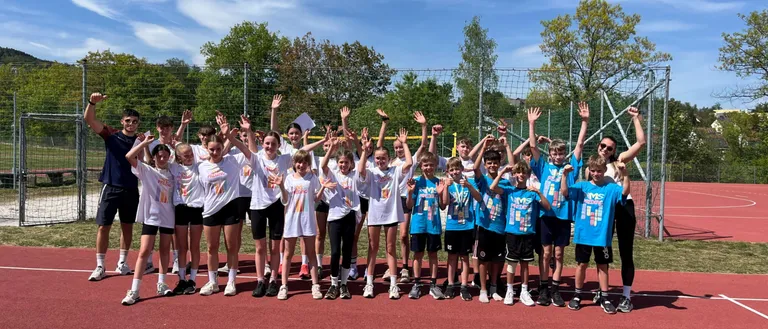 Eine Gruppe von Kindern in Sportuniform posiert auf einer Laufbahn, einige halten Medaillen. Sie sind von Bäumen und einem Zaun umgeben.