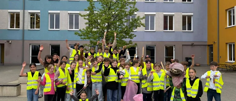 Eine große Gruppe von Kindern in Warnwesten posiert für ein Foto vor einem Gebäude. Viele halten Stöcke und Taschen. Ein Baum steht in der Mitte.