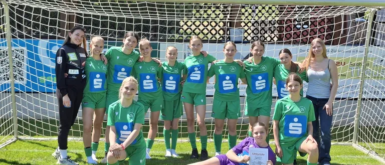 Eine Fußballmannschaft in grünen Trikots posiert für ein Foto vor einem Tor, wobei eine Spielerin ein Zertifikat in der Hand hält.
