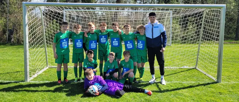 Eine Fußballmannschaft junger Jungen in grünen Trikots posiert vor einem Tor mit ihrem Trainer an einem sonnigen Tag.