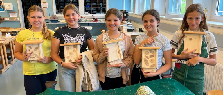 Fünf Mädchen posieren in einer Werkstatt und halten Holzvogelhäuschen. Der Raum ist hell beleuchtet, hat Fenster und einen grünen Tisch. Holzwerkzeuge und Materialien liegen auf dem Tisch.