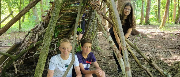 Drei Kinder sitzen auf einer Holzbank im Wald. Der Junge links trägt ein weißes Shirt. Der Junge in der Mitte trägt ein blaues Shirt. Eine Mädchen steht hinter ihnen.