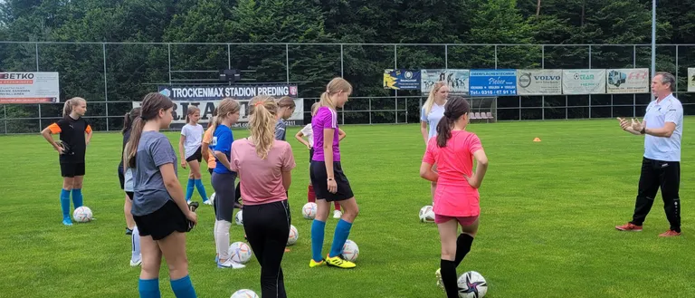 Eine Gruppe junger Mädchen trainiert Fußball auf einem Feld mit mehreren Bällen und Bäumen im Hintergrund.