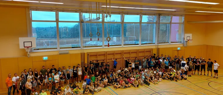 Eine Gruppe von Menschen versammelt sich auf einem Basketballplatz, einige stehend und andere sitzend, mit großen Fenstern und Blick auf Bäume draußen.