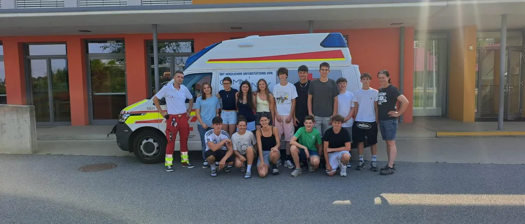 Eine Gruppe junger Menschen und Erwachsener posiert für ein Foto vor einem Krankenwagen. Der Krankenwagen ist vor einem Gebäude mit Glasfenstern und einer Tür geparkt.