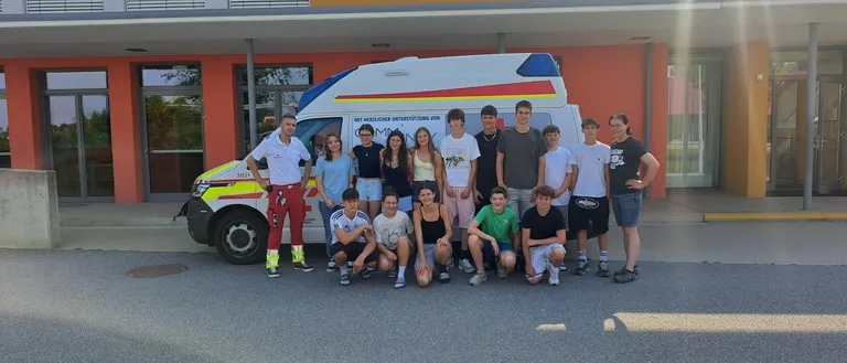 Eine Gruppe junger Menschen und Erwachsener posiert für ein Foto vor einem Krankenwagen. Der Krankenwagen ist vor einem Gebäude mit Glasfenstern und einer Tür geparkt.