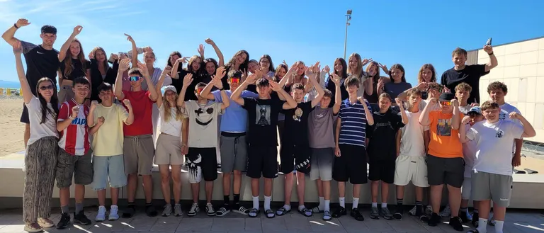 Eine Gruppe junger Menschen posiert an einem sonnigen Tag für ein Foto. Sie stehen in zwei Reihen auf einem gefliesten Boden, manche mit erhobenen Händen. Hinter ihnen ist ein klarer blauer Himmel.
