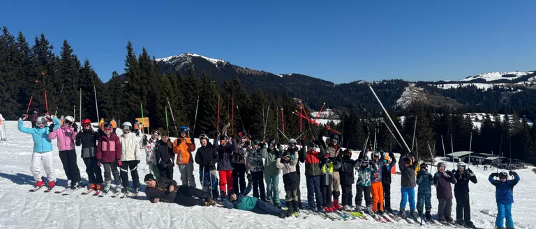 Eine Gruppe von Skifahrern und Snowboardern posiert für ein Foto auf einem verschneiten Berg mit Bäumen und Bergen im Hintergrund.
