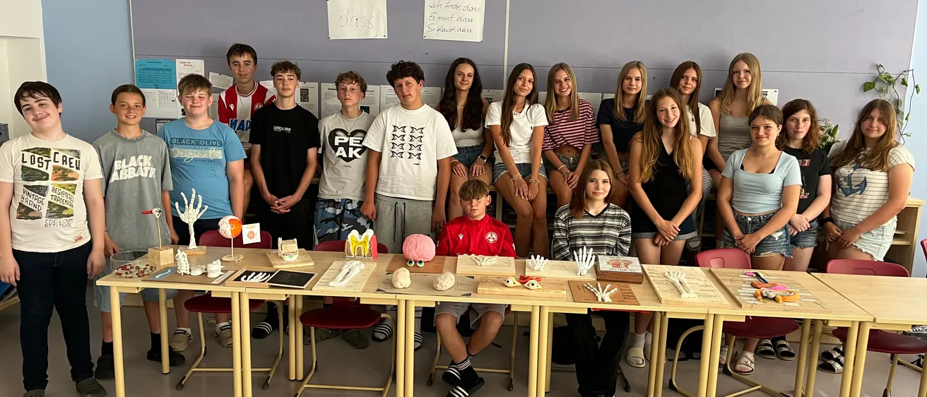 Eine Gruppe von Schülern posiert für ein Foto in einem Klassenzimmer mit Tischen voller anatomischer Modelle. Die Schüler sitzen und stehen, einige tragen Turnschuhe und andere lässige Kleidung.