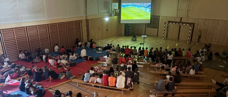 Eine Gruppe von Menschen sitzt auf Bänken und schaut sich ein Fußballspiel auf einer großen Leinwand in einer Turnhalle an.