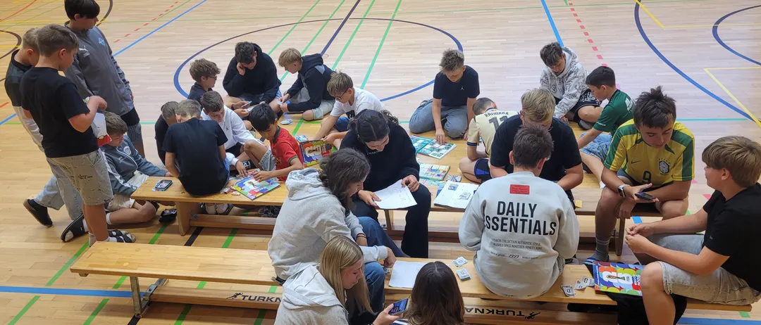 Schüler sitzen auf Bänken in einer Turnhalle, einige mit Büchern, andere am Telefon. Eine Person mit einem Daily Essentials Sweatshirt sitzt vorne. Bunte Linien kennzeichnen den Platz.