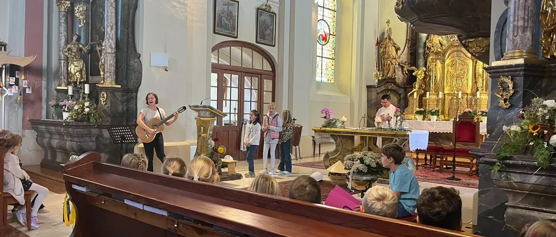 Eine Frau spielt Gitarre in einer Kirche, während eine Gruppe von Kindern und Erwachsenen zuschaut.