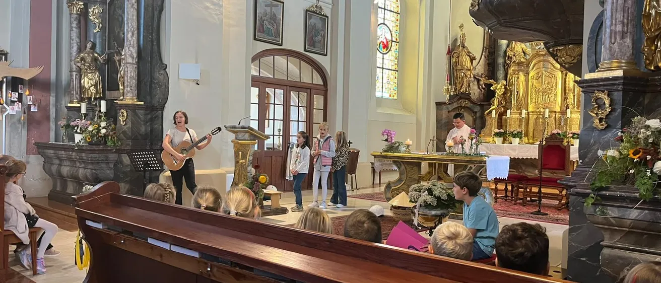 Eine Frau spielt Gitarre in einer Kirche, während eine Gruppe von Kindern und Erwachsenen zuschaut.