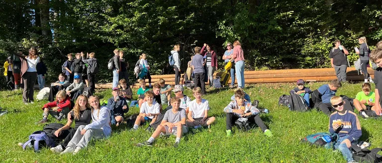 Eine Gruppe von Menschen sitzt auf einem Grasfeld, einige stehen und andere sitzen auf dem Boden. Sie sind wahrscheinlich Schüler.