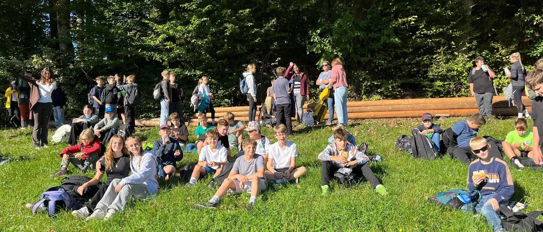 Eine Gruppe von Menschen sitzt auf einem Grasfeld, einige stehen und andere sitzen auf dem Boden. Sie sind wahrscheinlich Schüler.
