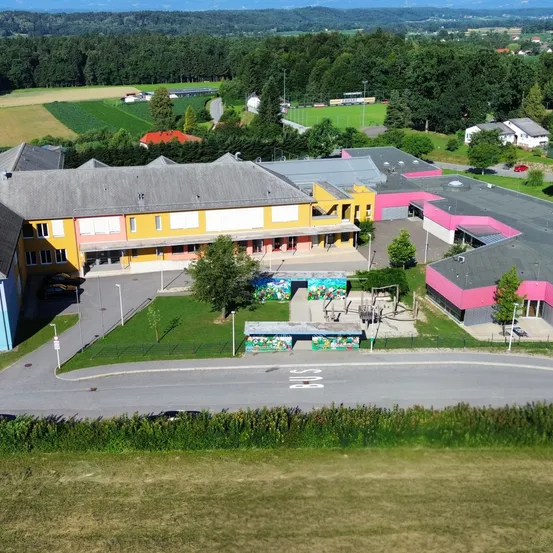 Luftaufnahme einer Schule mit gelben und rosa Gebäuden, umgeben von Bäumen und Gras, mit einem Spielplatz und einem Parkplatz davor.