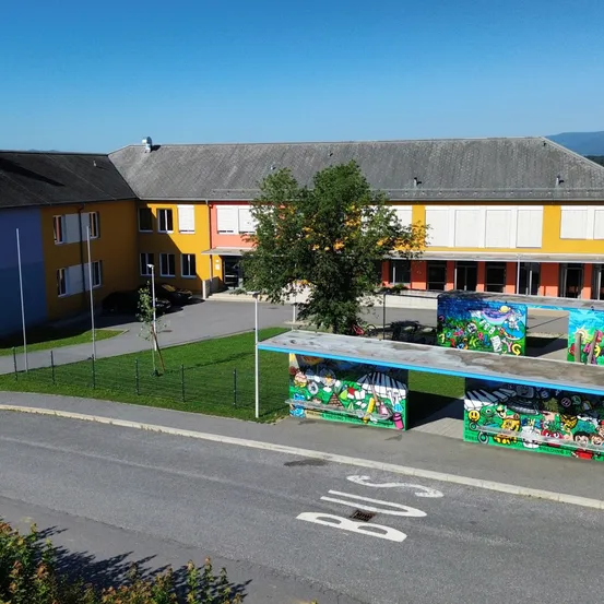 Luftaufnahme eines farbenfrohen Schulgebäudes mit einem Baum und einer Bushaltestelle davor, unter einem klaren blauen Himmel.