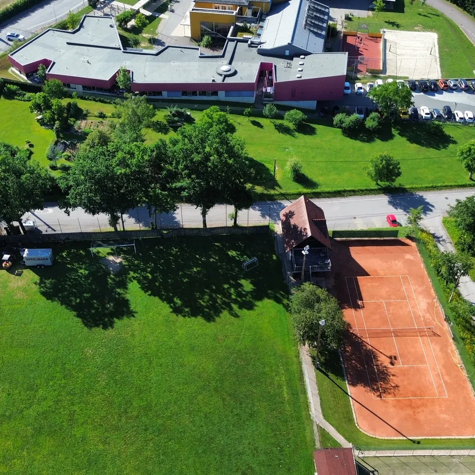 Eine Luftaufnahme einer Sportanlage mit mehreren Gebäuden und Tennisplätzen. Das Hauptgebäude ist pink und grau. Das Gelände hat grünen Rasen und Bäume. Autos sind auf dem Parkplatz geparkt.