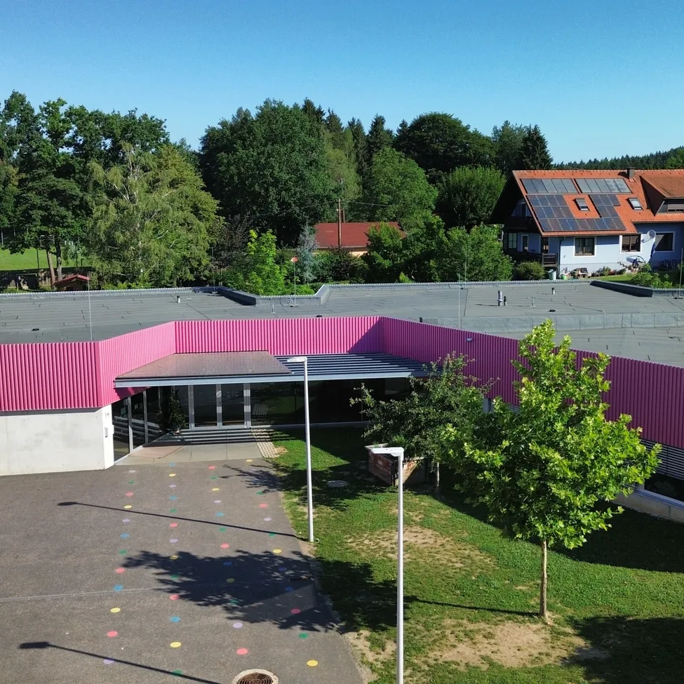 Luftaufnahme einer Schule mit rosa Fassade und grauem Dach. Die Schule ist von einem Spielplatz mit bunten Punkten umgeben. Im Hintergrund stehen Bäume und Häuser, eines davon mit Solarmodulen.