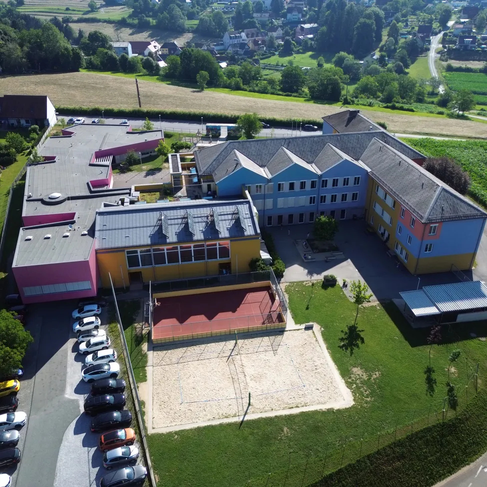Ein Luftbild eines mehrfarbigen Gebäudes mit Tennisplatz, Sandgrube und Parkplatz. Das Gebäude hat mehrere Fenster, und viele Autos sind draußen geparkt. Das Gebiet ist von grünem Gras und Bäumen umgeben.
