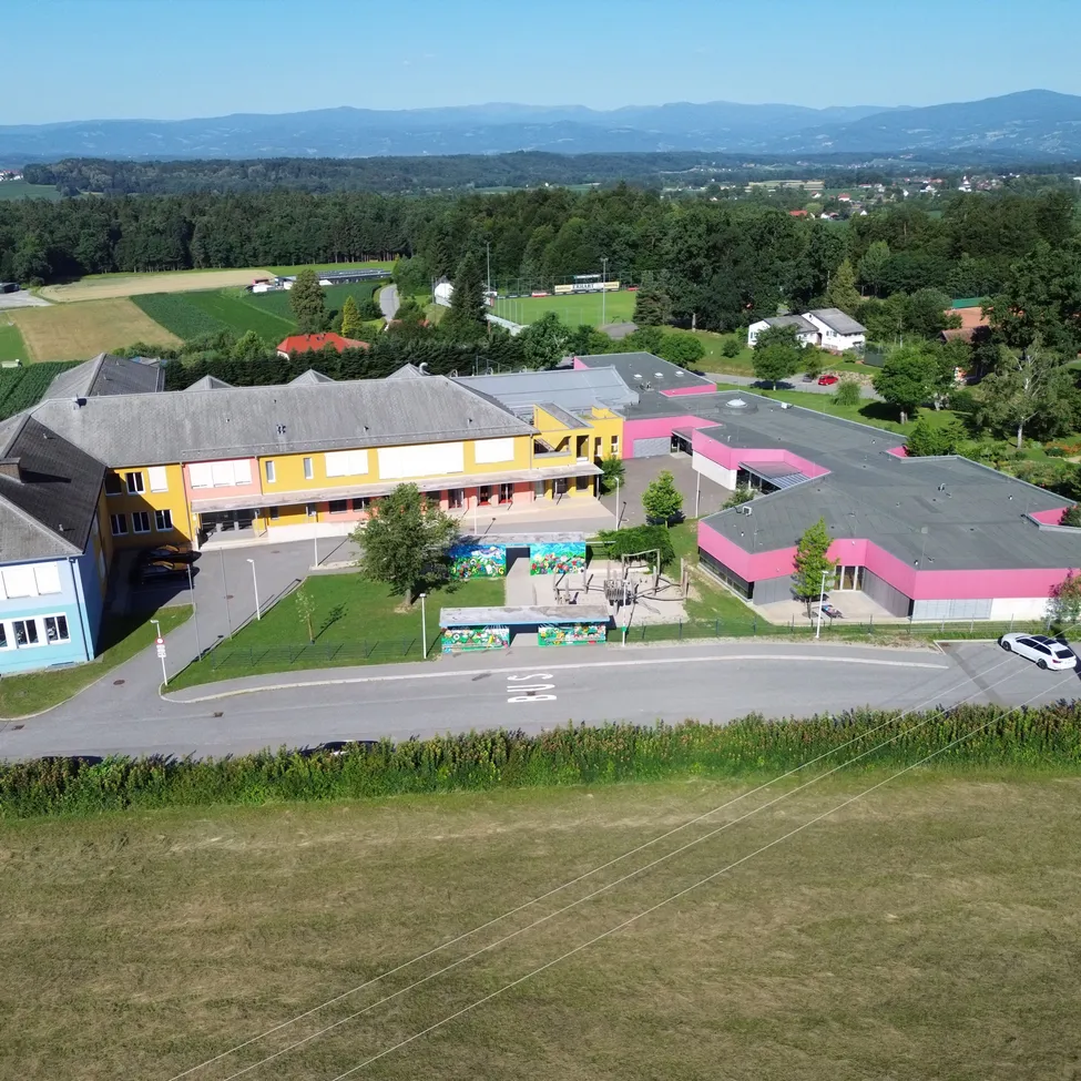 Luftaufnahme einer Schule mit gelben und pinken Wänden, in einem ländlichen Gebiet gelegen. Die Schule besteht aus mehreren Gebäuden und ist von viel Grün umgeben. Ein Spielplatz ist in der Nähe der Straße zu sehen.