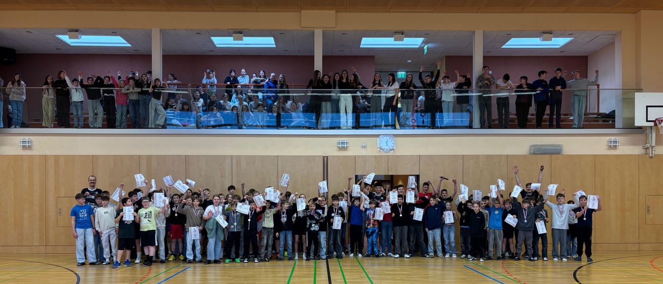 Eine Gruppe von Kindern und Jugendlichen feiert auf einem Basketballplatz, während viele Zuschauer von der Galerie zuschauen. Alle halten Urkunden hoch.