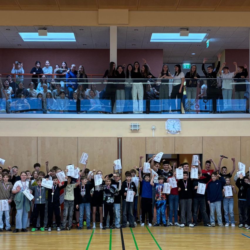 Eine Gruppe von Kindern und Jugendlichen feiert auf einem Basketballplatz, während viele Zuschauer von der Galerie zuschauen. Alle halten Urkunden hoch.