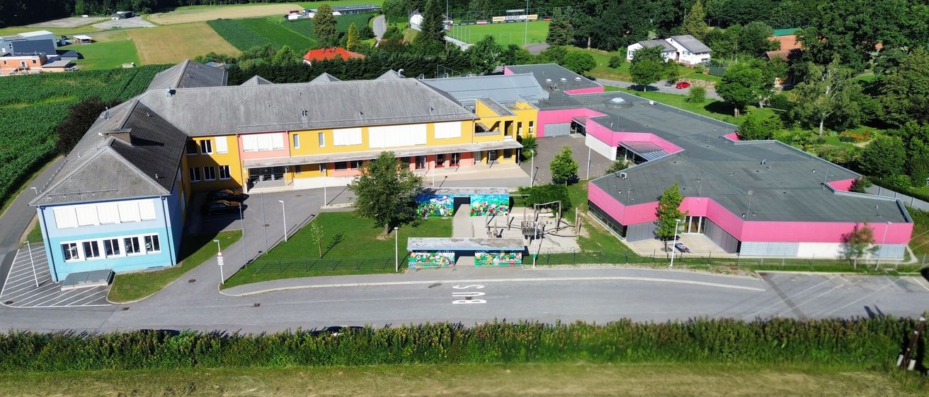 Ein Luftbild einer Schule mit gelben und rosa Gebäuden, umgeben von Gras, Bäumen und einer Straße. Vor dem Gebäude sind Autos geparkt, und in der Nähe befindet sich eine Bushaltestelle. In der Ferne stehen Häuser und ein Feld.