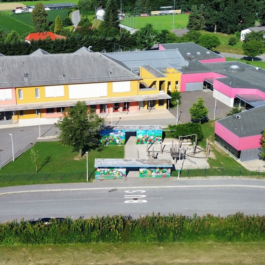 Ein Luftbild einer Schule mit gelben und rosa Gebäuden, umgeben von Gras, Bäumen und einer Straße. Vor dem Gebäude sind Autos geparkt, und in der Nähe befindet sich eine Bushaltestelle. In der Ferne stehen Häuser und ein Feld.