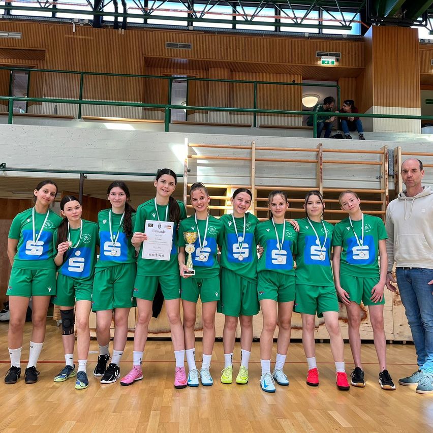 Eine Gruppe von Sportlerinnen in grünen Uniformen mit Medaillen um den Hals, die einen Pokal und ein Zertifikat halten, steht zusammen mit einem Mann in einer Turnhalle.