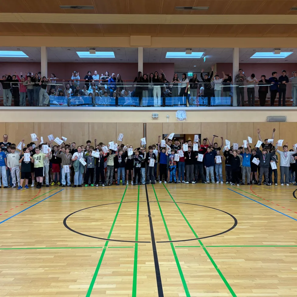 Eine große Gruppe von Menschen, wahrscheinlich Schüler, versammelt sich in einer Turnhalle und hält Urkunden in der Hand. Zuschauer jubeln auf der Galerie darüber. Der Hallenboden hat Basketball-Markierungen.