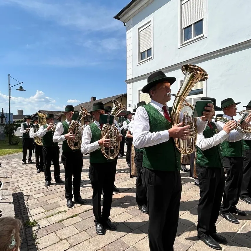 Eine Gruppe von Männern in grünen Westen und schwarzen Hüten spielt Blechblasinstrumente vor einem weißen Gebäude.