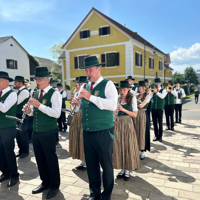 Eine Gruppe von Musikern in grünen Westen und schwarzen Hosen spielt im Freien. Sie halten verschiedene Musikinstrumente wie Trompeten, Flöten und Posaunen. Die Musiker stehen vor einem gelben Gebäude mit mehreren Fenstern.