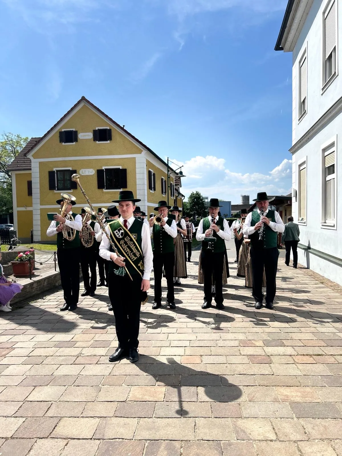 Eine Blaskapelle in grünen Uniformen, schwarzen Hüten und Schuhen tritt vor einem gelben Haus an einem sonnigen Tag auf.