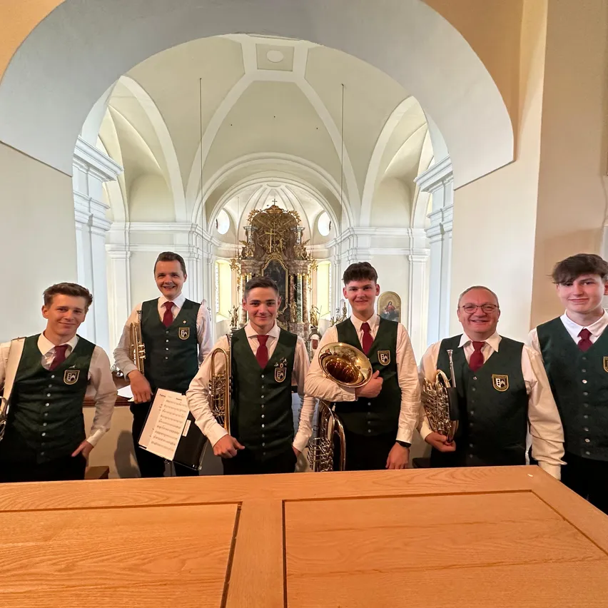 Sechs junge Männer in grünen Westen mit weißen Hemden und roten Krawatten stehen in einer Kirche und posieren für ein Foto mit Musikinstrumenten.