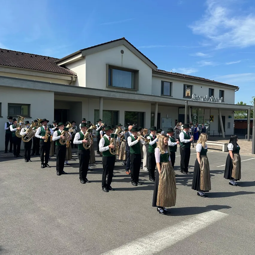 Ein Blasorchester in traditioneller deutscher Kleidung spielt im Freien vor einem Gebäude mit der Aufschrift 'Genossenschaft'.