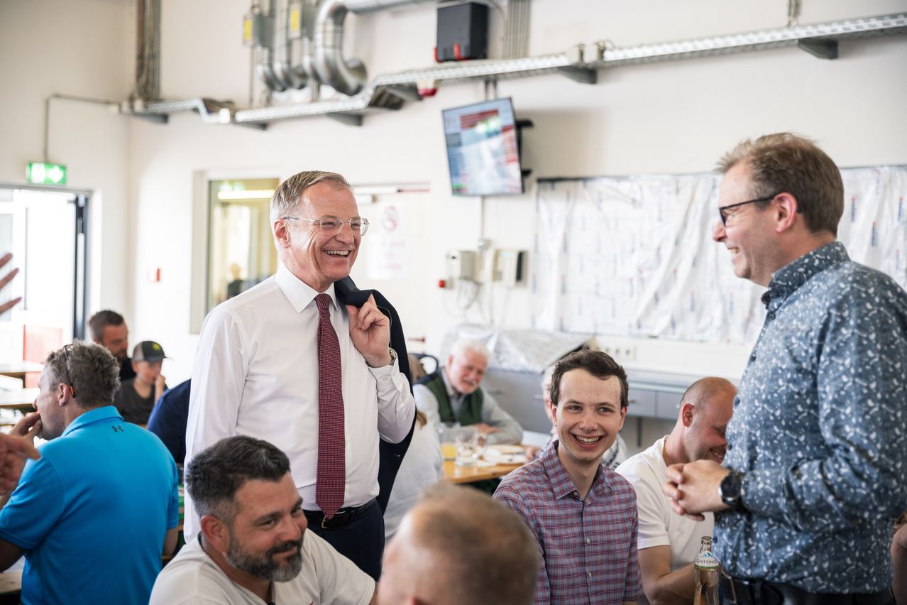 Eine Gruppe von Menschen ist in einer Inneneinrichtung versammelt. Ein älterer Mann in einem weißen Hemd und einer Krawatte lächelt. Ein junger Mann in einem karierten Hemd lacht. Es gibt einen Tisch mit Getränken und einen Fernseher an der Wand.